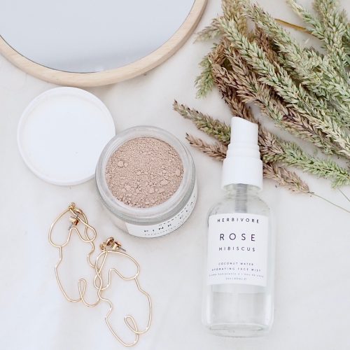 Flat lay photo showing herbivore botanicals face spray and rose face mask, also contains dry grass, monki woman's body silhouette drop earrings and wooden mirror from H&M.