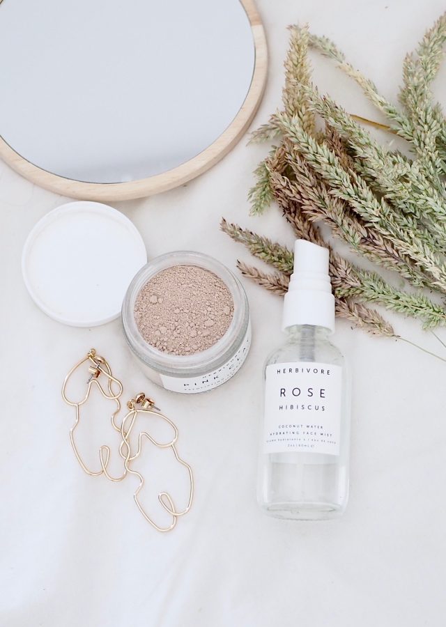Flat lay photo showing herbivore botanicals face spray and rose face mask, also contains dry grass, monki woman's body silhouette drop earrings and wooden mirror from H&M.
