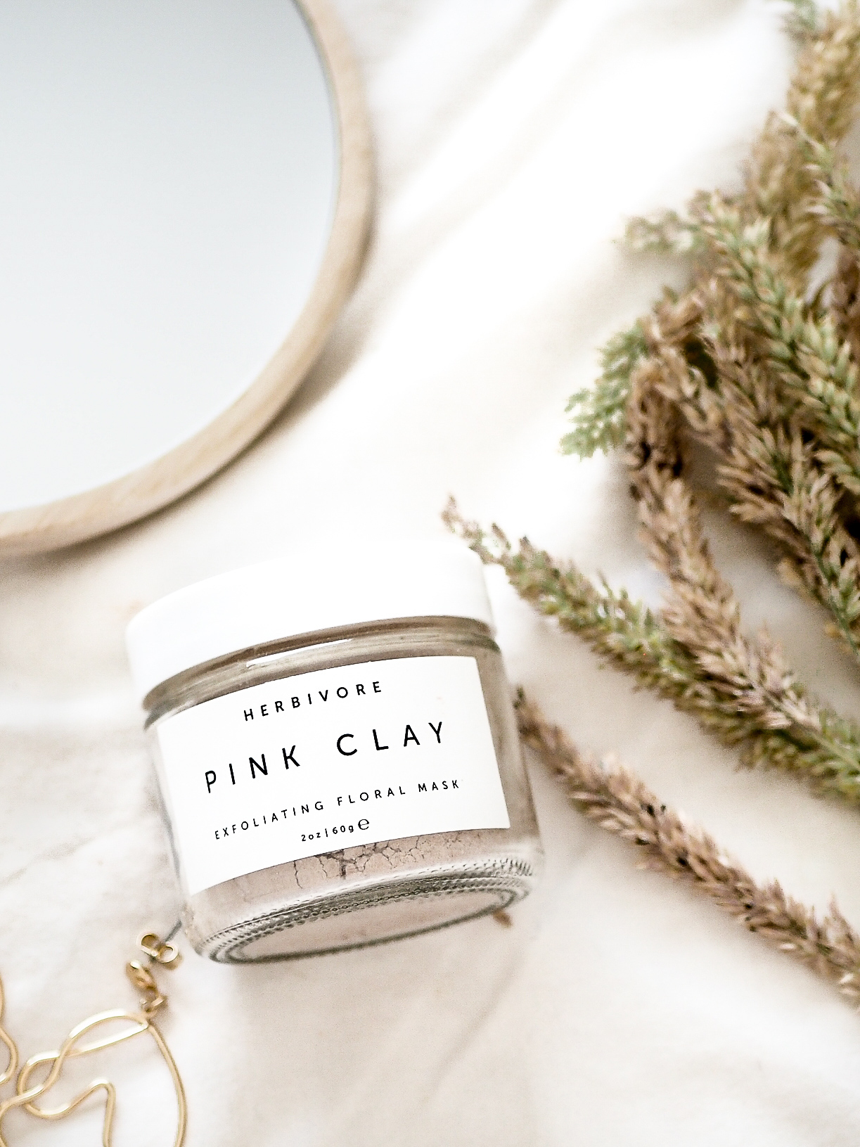 flatlay of herbivore botanicals pink clay mask with dried grass and wooden mirror