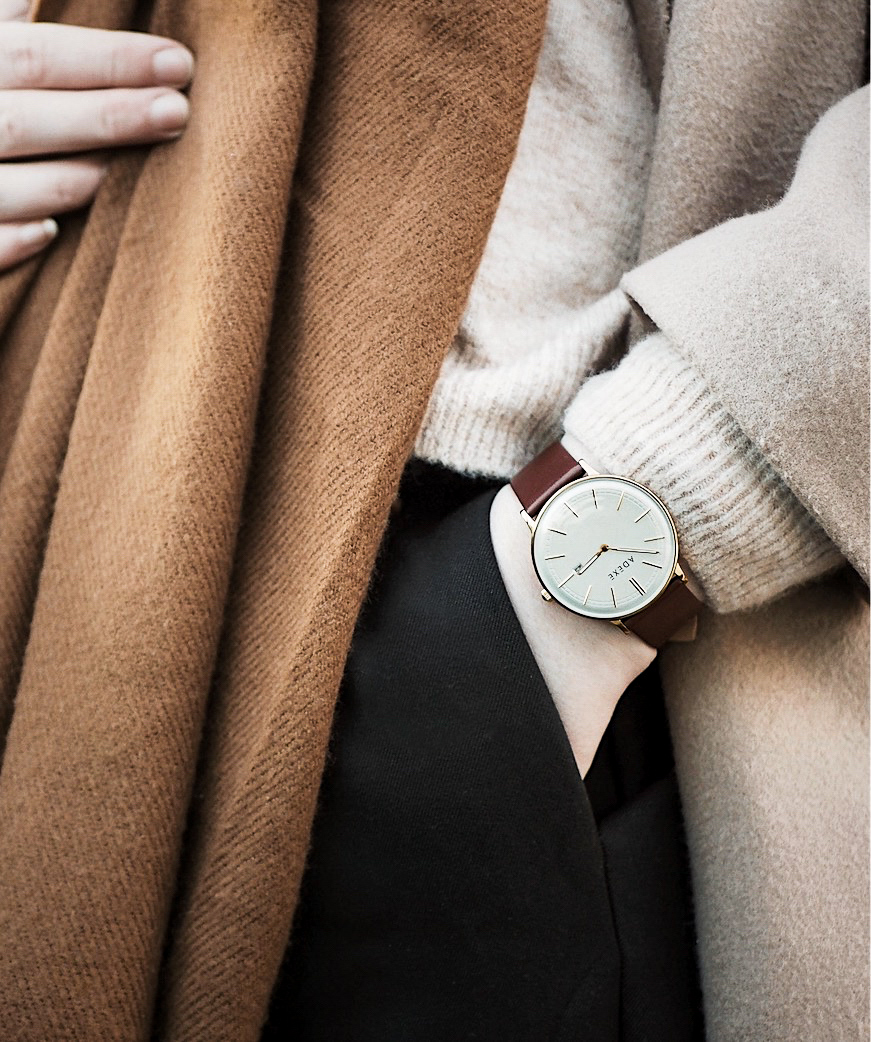 androgynous styling of adexe meek watch, with hand in trousers pocket and beige jumper, brown scarf and beige winter coat